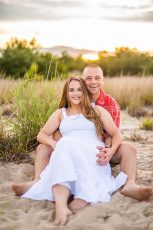 Sunset beach portrait of couple in white dress and red plaid shirt sitting on sand.