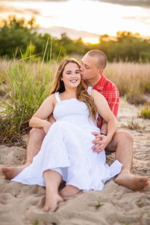 Romantic couple sitting on sandy beach at sunset wearing white dress and red plaid shirt.