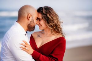 Intimate couple moment on beach with waves in background and matching red and white outfits.
