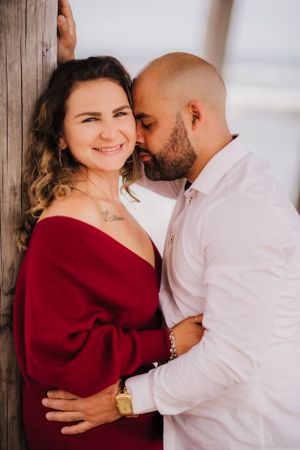 Romantic couple embracing against wooden wall at beach with red sweater and white shirt.