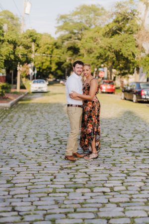 A couple poses together on a cobblestone street with parked cars and trees in the background.