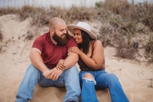 A relaxed couple sitting together on sandy dunes during their outdoor photoshoot.