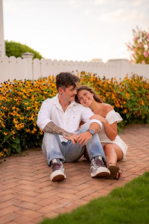 A couple sits together on brick pavement surrounded by blooming yellow flowers and a white fence.