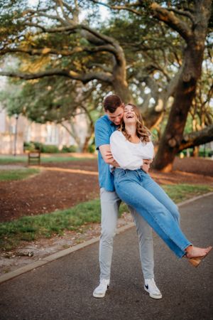 Playful couple dancing and laughing under large oak trees in a park setting.