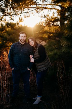 A couple poses together in front of a golden sunset through pine trees.