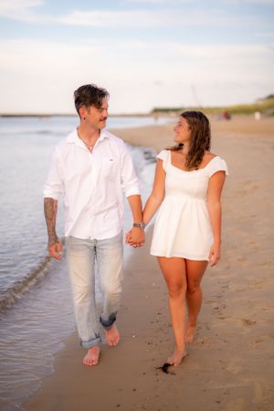 A couple walks hand in hand along the beach shoreline at sunset in casual white outfits.