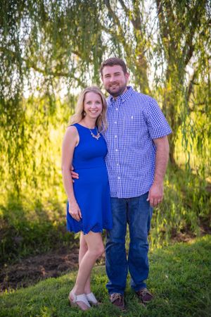 Couple in blue dress and checkered shirt pose together under willow tree.
