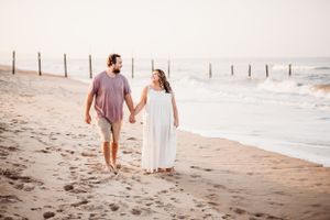 Couple walking together on sandy beach with fence posts and ocean waves in background.