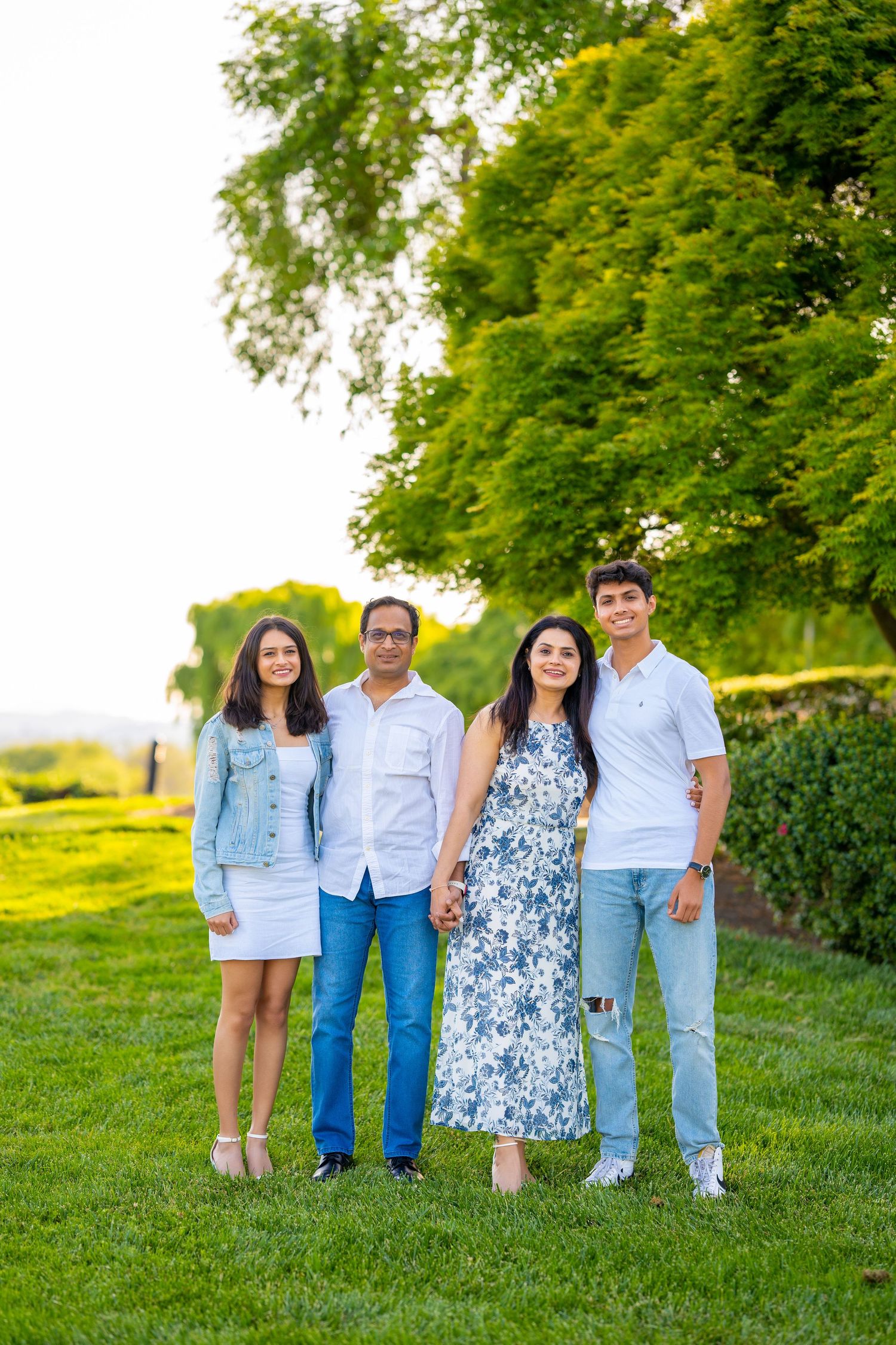 Family of four poses outdoors on grassy hill with green trees in casual summer attire.