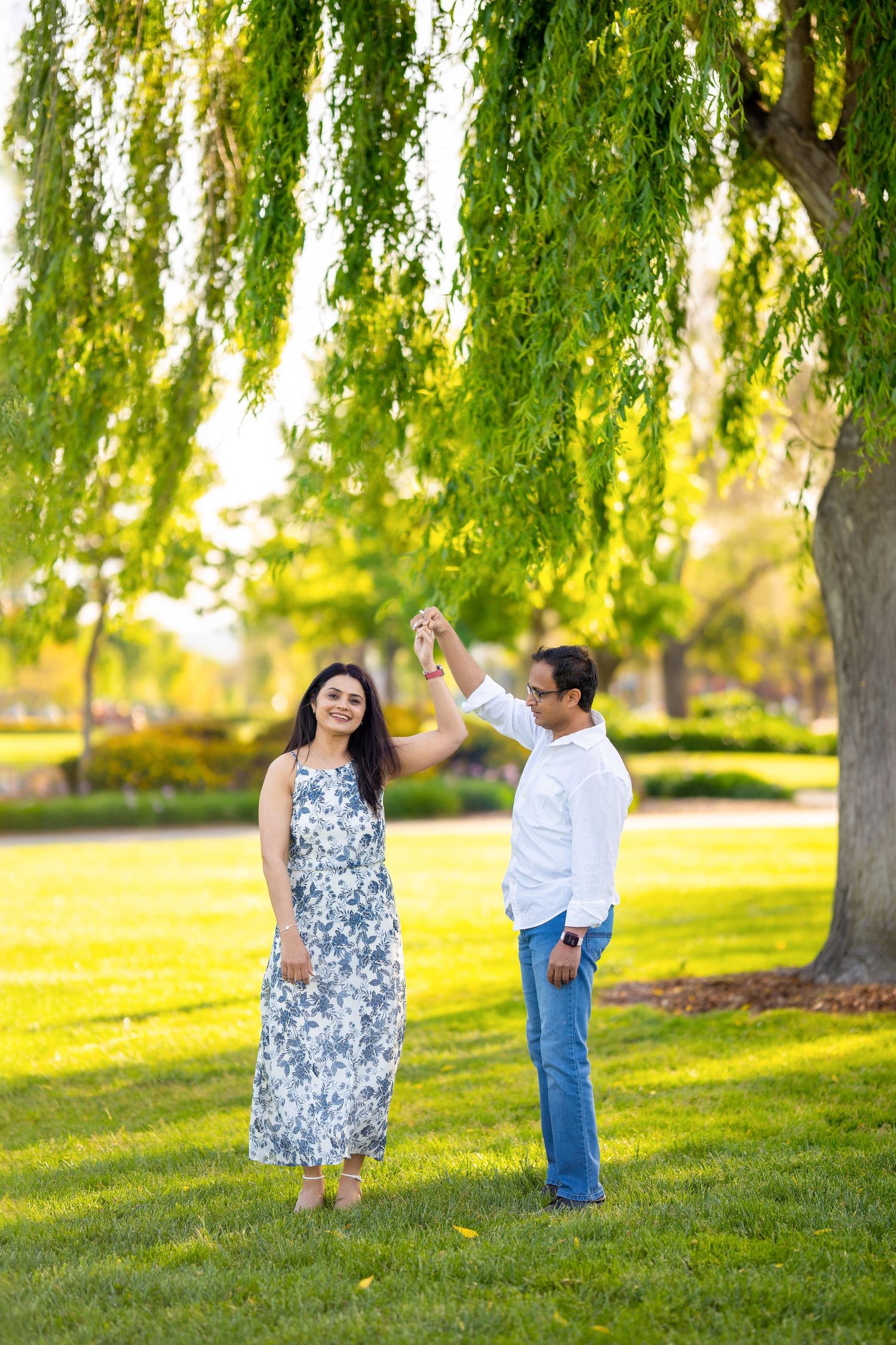Couple poses under weeping willow tree in park during sunset photoshoot.