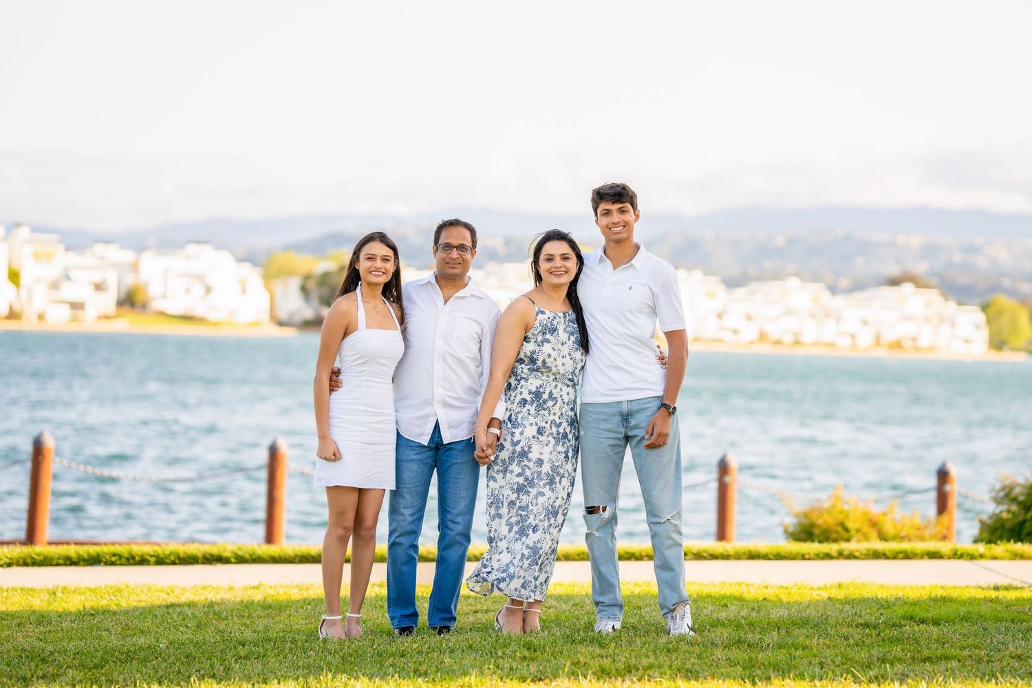 Family smiles together on waterfront lawn with coastal scenery in background.