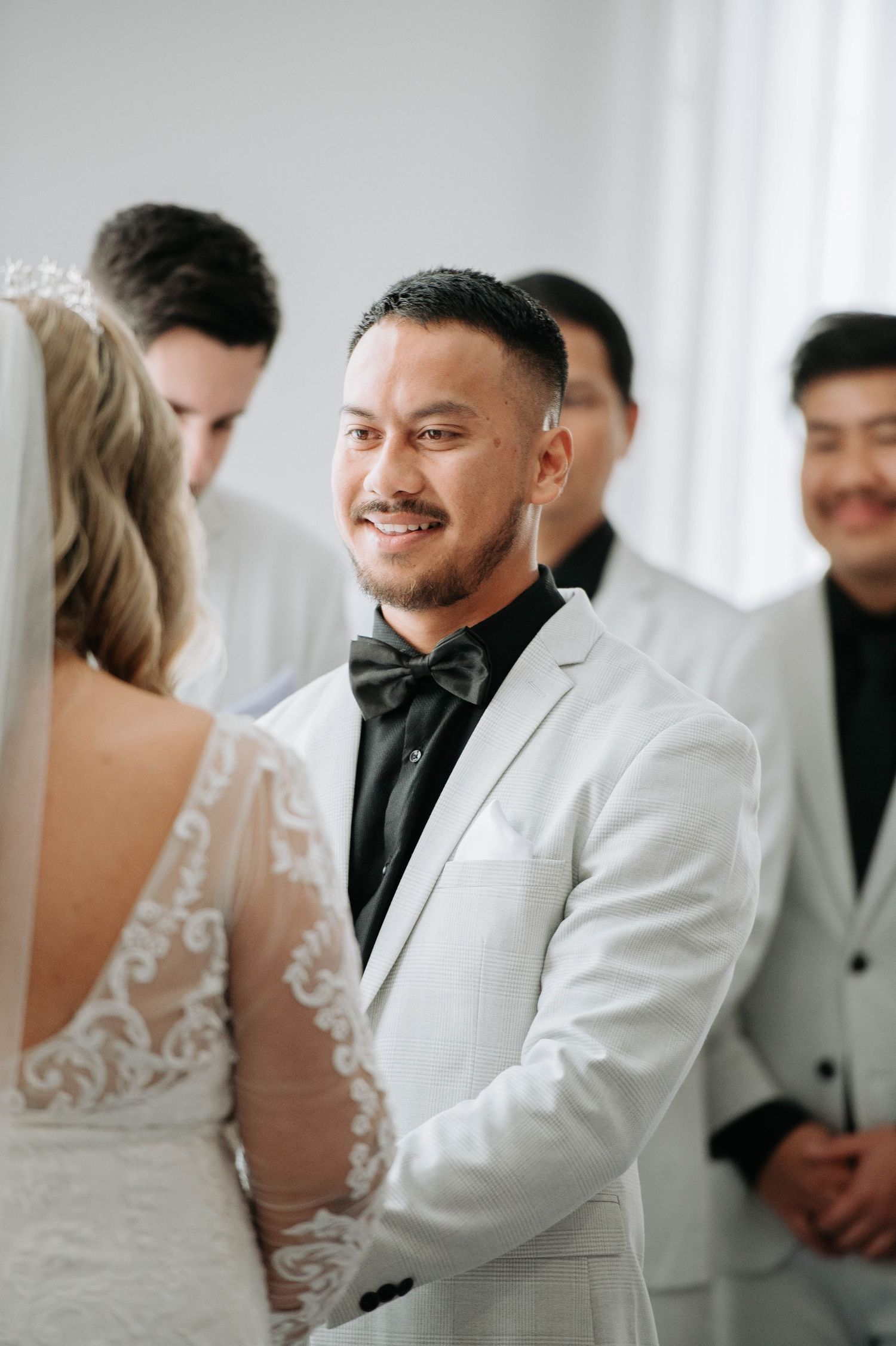 Groom in light grey suit and bow tie smiles during wedding ceremony.