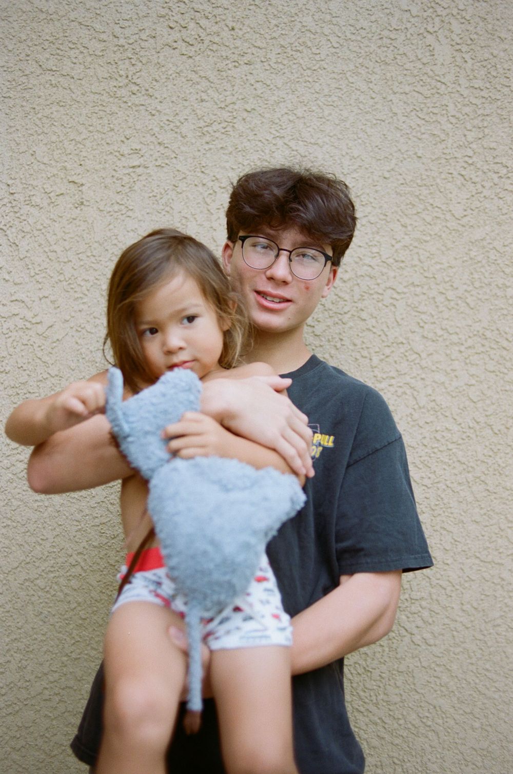 Two people embracing while holding a blue stuffed animal against a beige wall.