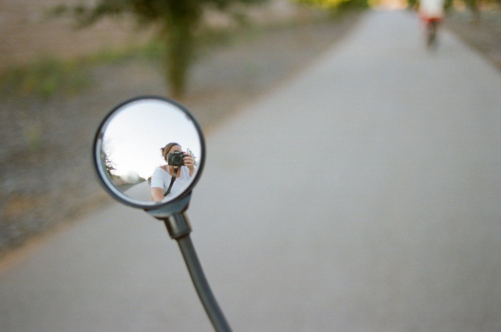 Bike mirror reflection showing a path and distant figure on a sunny day.