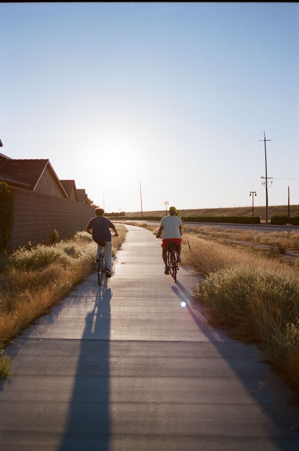 Two cyclists ride along a paved path at sunset casting long shadows beside a residential area.