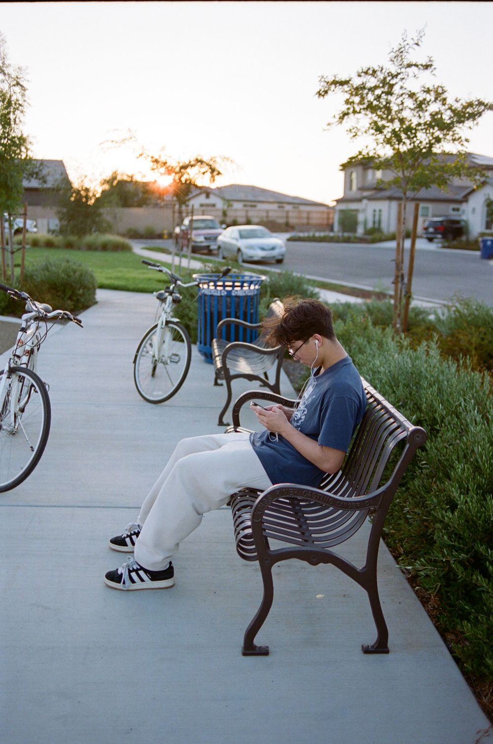 Person sitting on a park bench looking at phone with bicycles parked nearby at sunset.