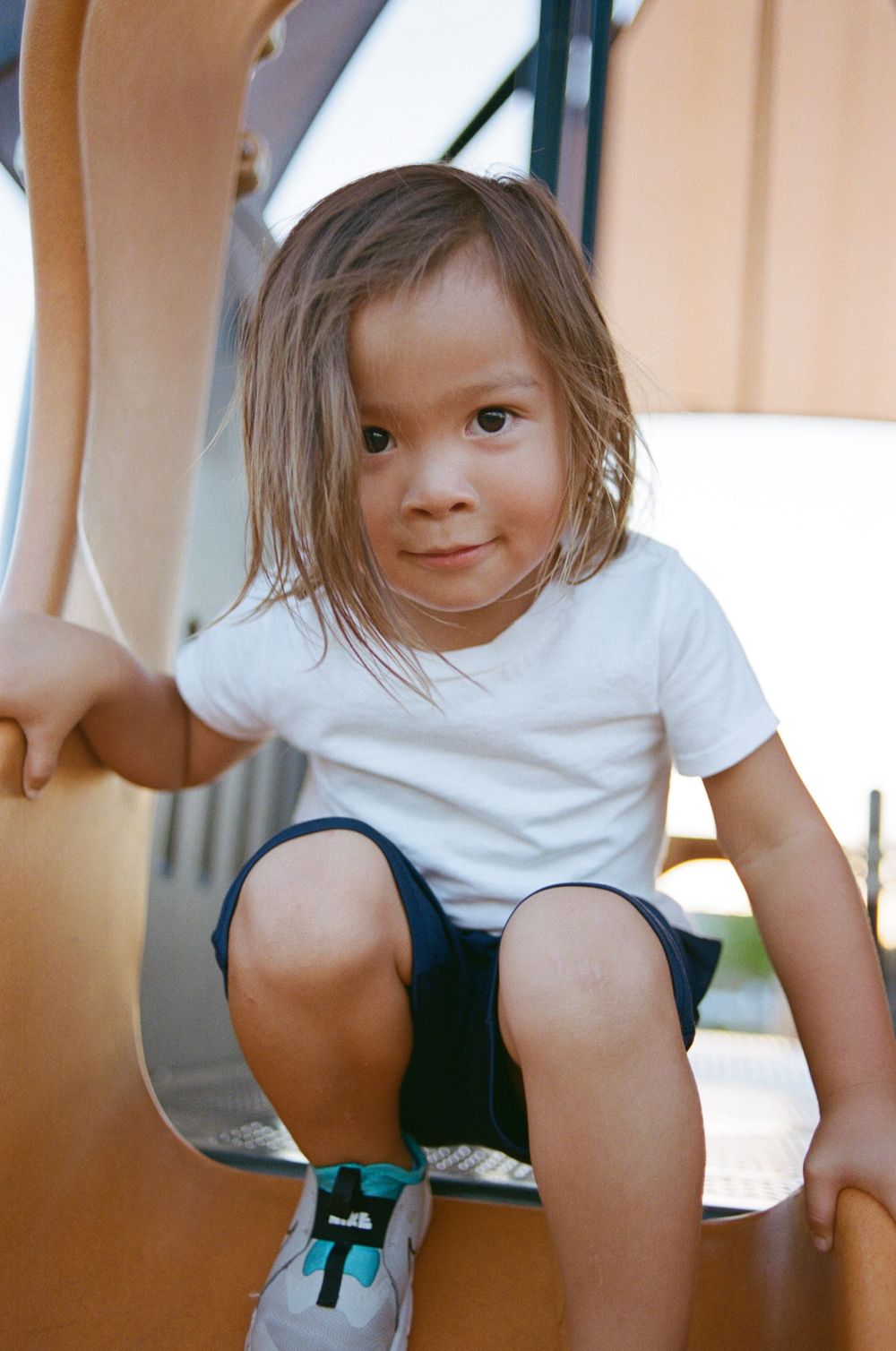 Young child in white t-shirt and shorts sitting on playground equipment.