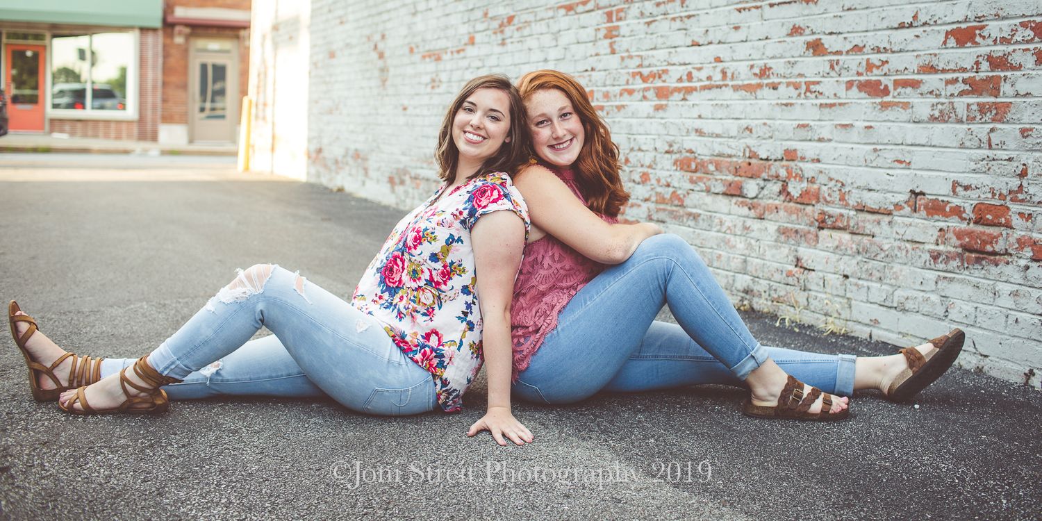 Two friends in casual outfits sit together against a rustic brick wall.