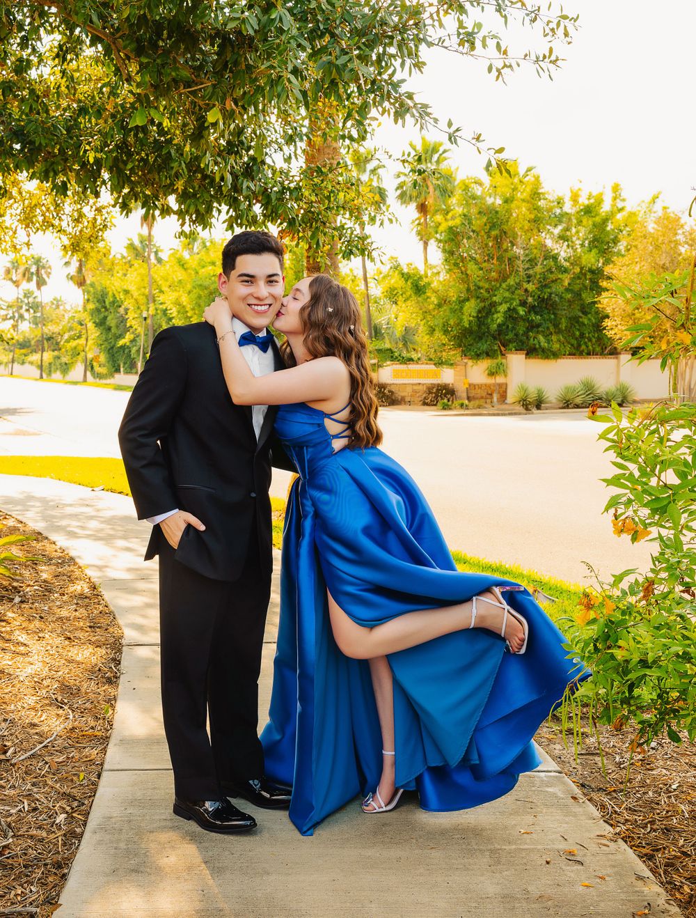 Playful formal photo of a couple on a garden path with her leg lifted in a flirty pose.