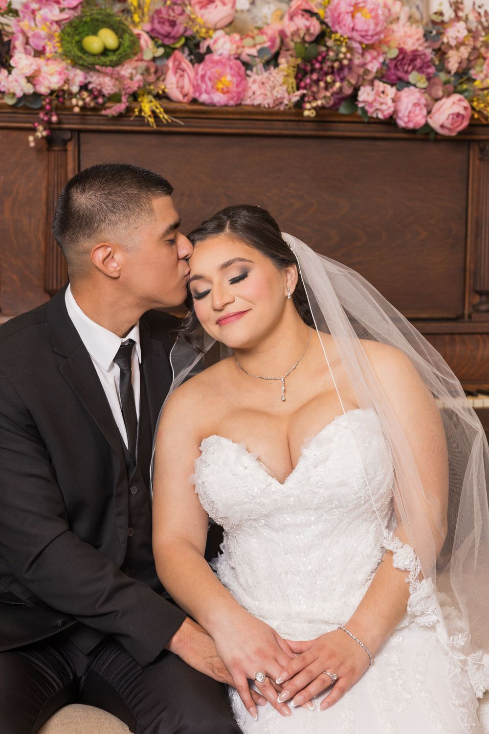 An intimate wedding portrait shows a couple posed by a piano with pink flowers.