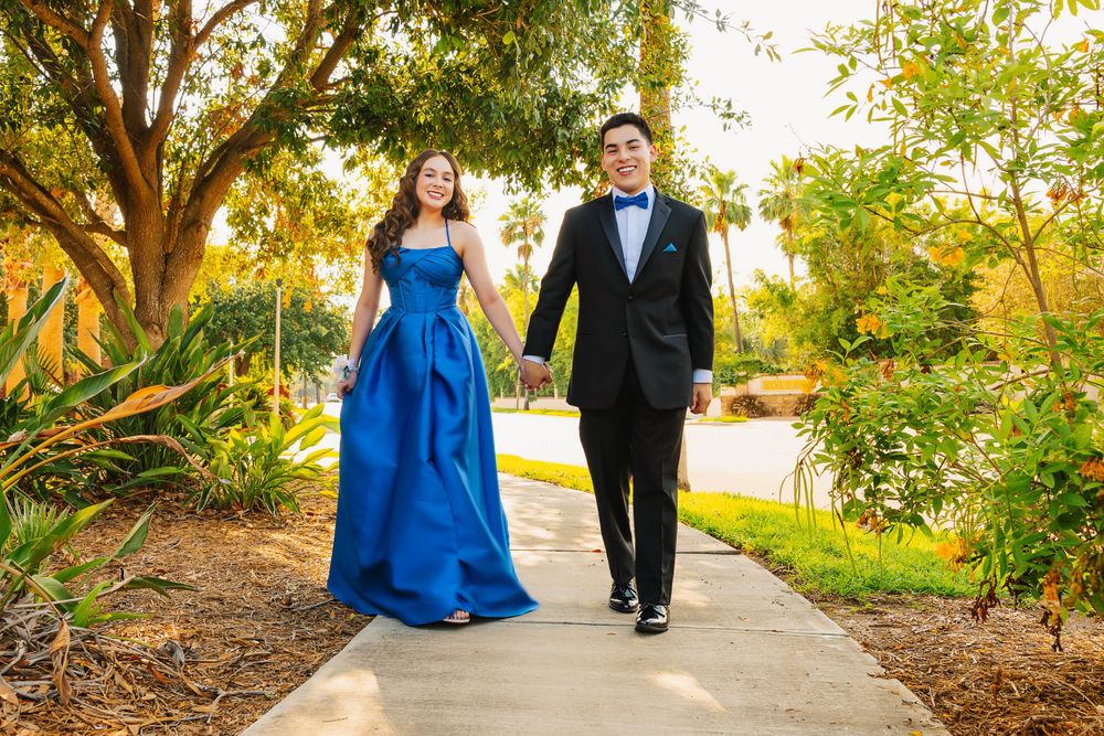 Couple in formal attire walks together on a garden path surrounded by lush greenery.