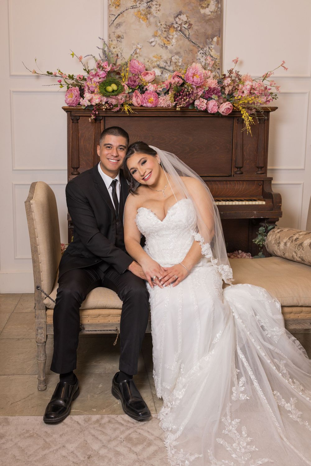 A bride in a white lace dress sits with her groom next to an upright piano decorated with pink floral arrangements.