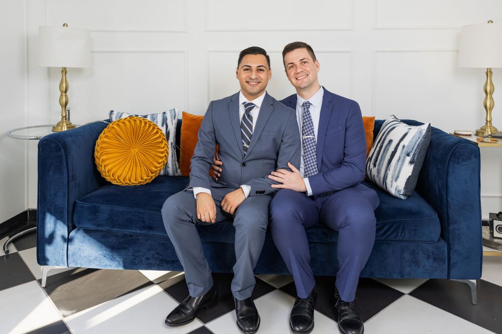 Two people in suits sit together on a navy blue velvet couch with decorative pillows.