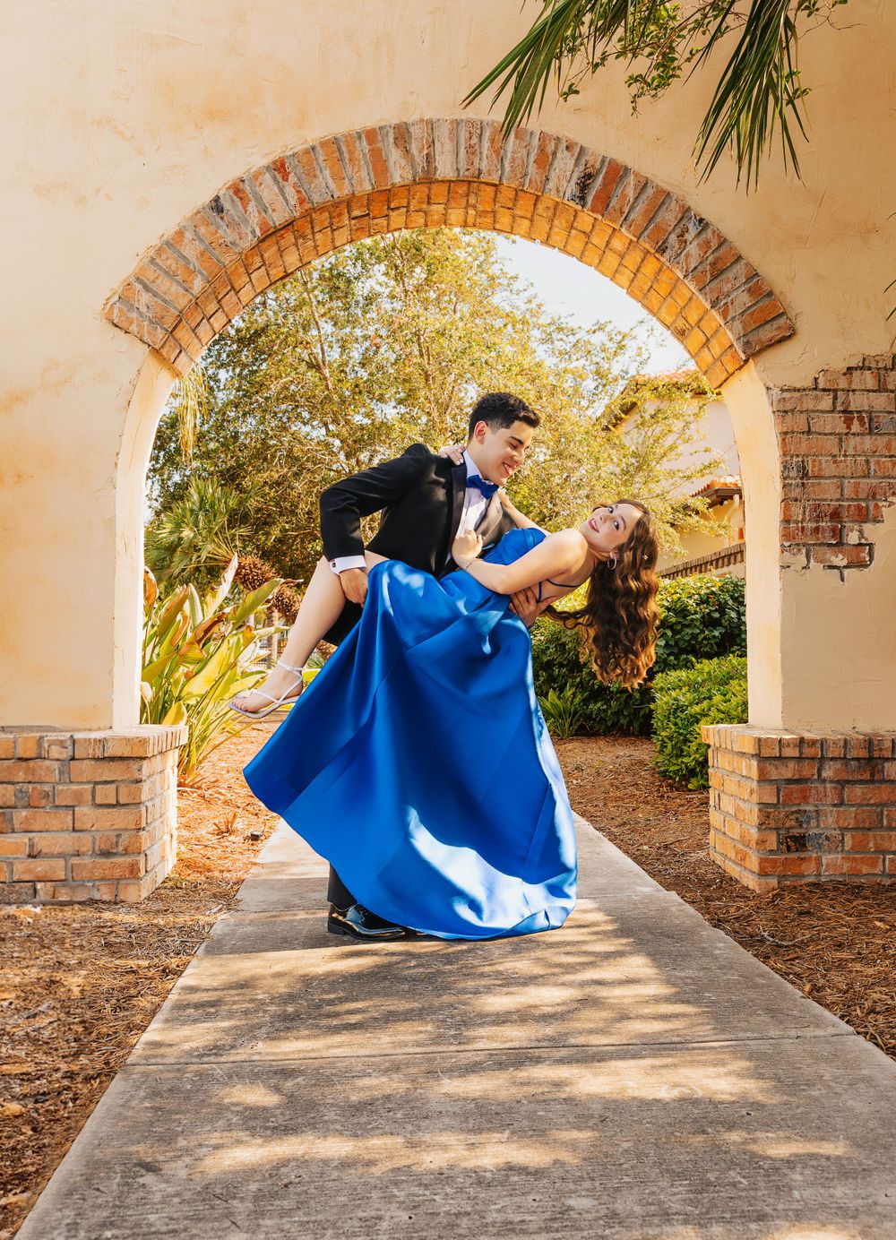 A romantic dip pose under a brick archway with a flowing blue ball gown and black tuxedo.