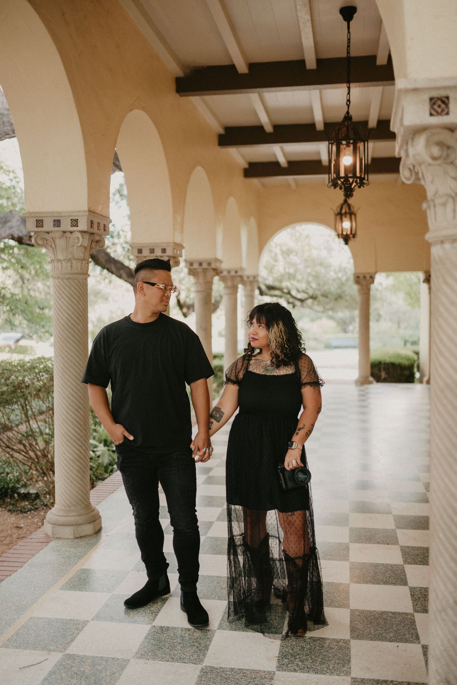 Stylish couple in black outfits walking under ornate Spanish colonial arches with checkered floor.