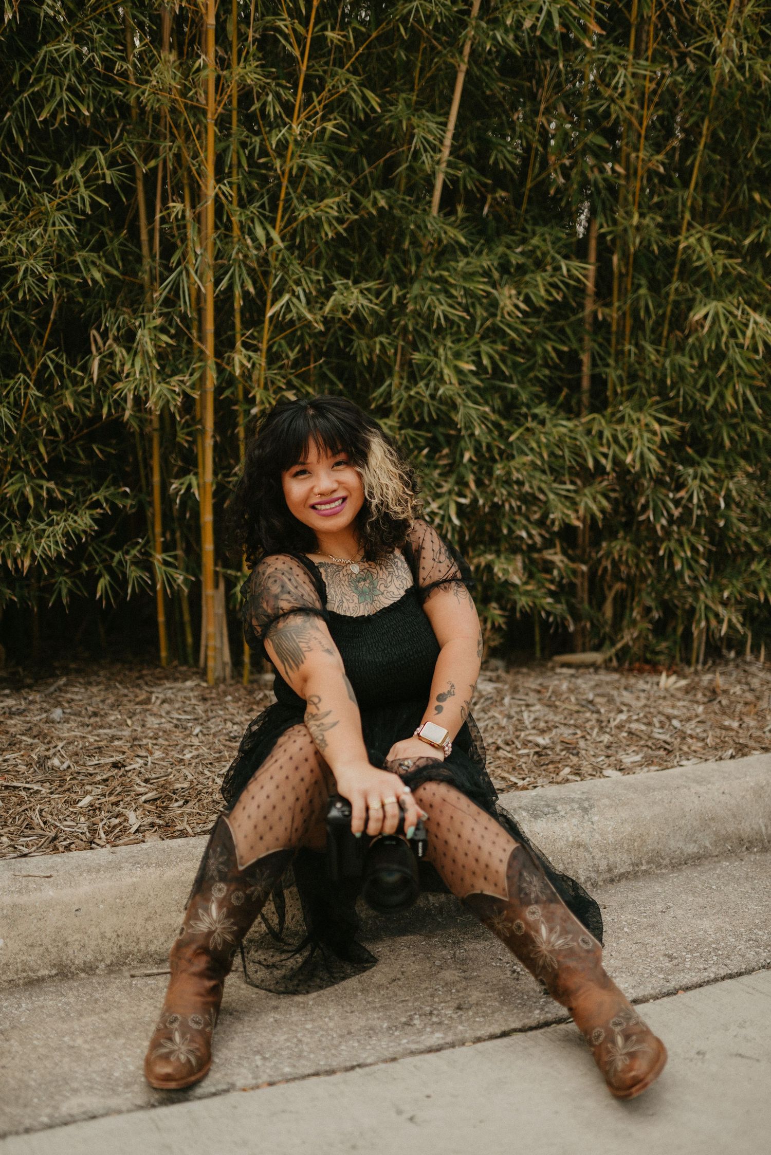 A photographer in a black dress and boots poses against a bamboo garden backdrop.