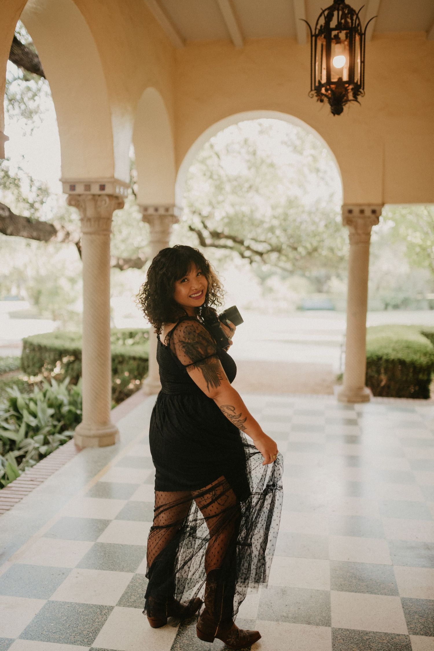 A photographer in a black lace dress poses in an elegant colonnade with checkerboard flooring and hanging lanterns.