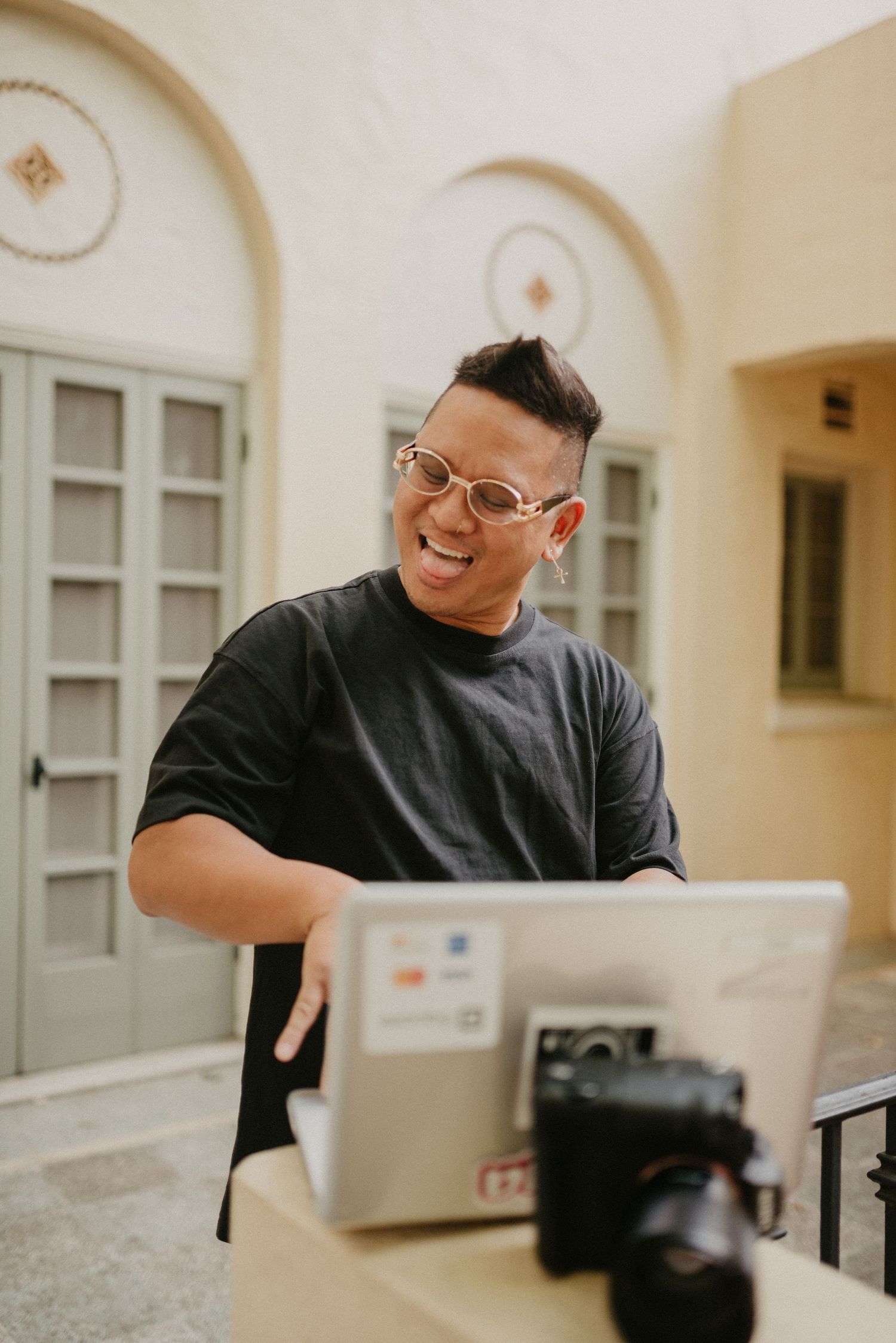 A photographer smiles while working on a laptop with camera equipment on an outdoor patio.