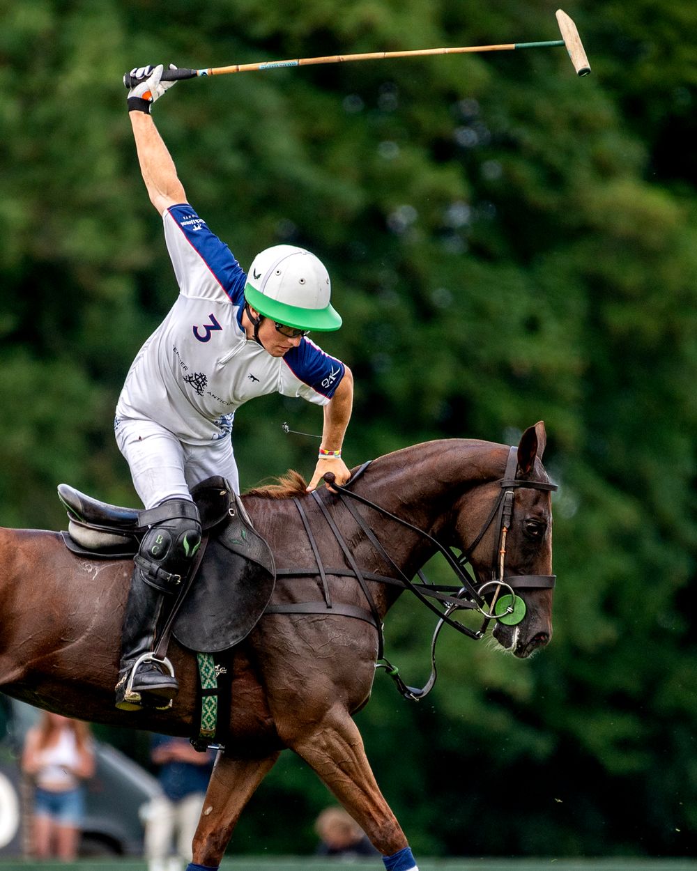 Polo player wearing a white No.3 jersey and green helmet striking the ball in a powerful offside swing while riding a dark bay pony.