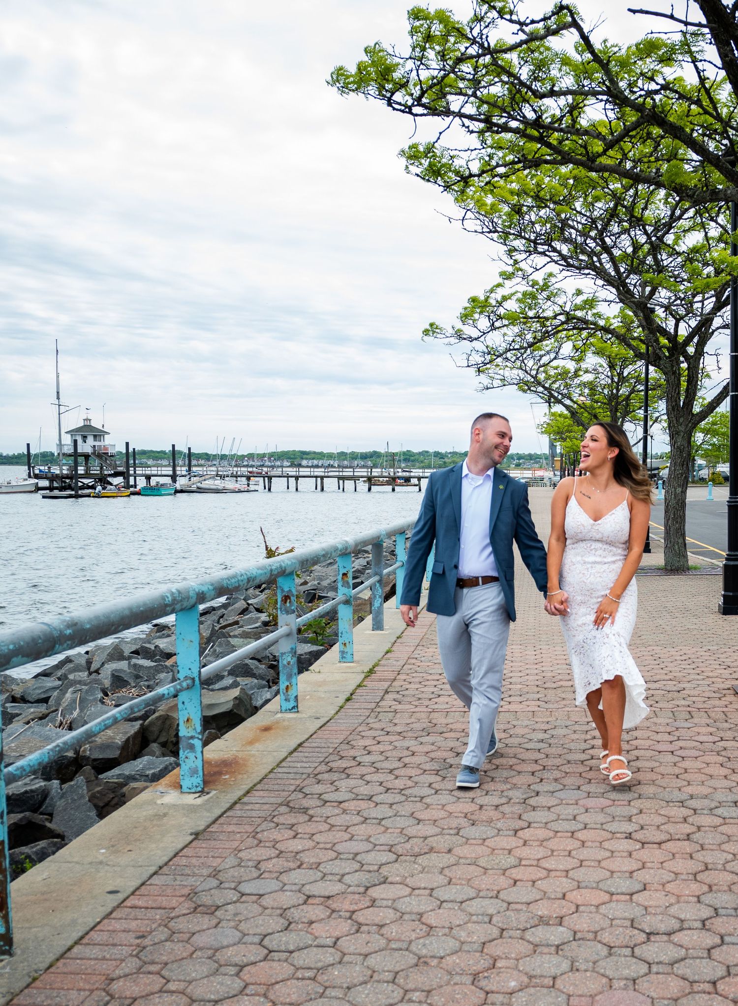 A newly married couple strolls along a waterfront promenade with marina docks and boats in the background on a sunny day.