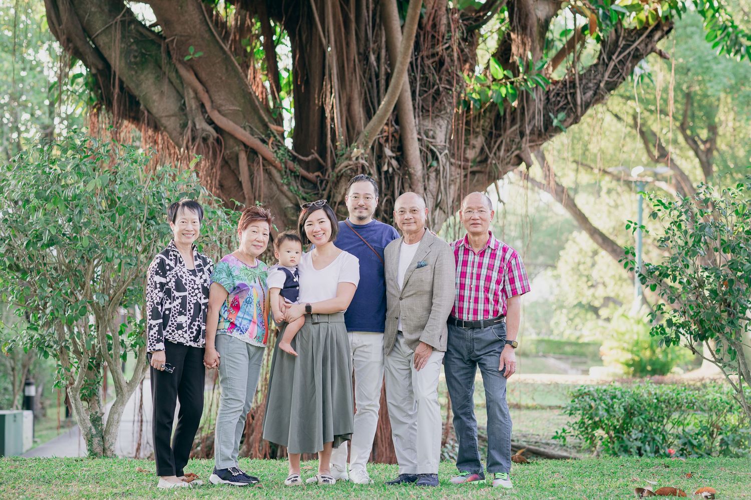 A cheerful group stands together under a large banyan tree in a lush garden setting.