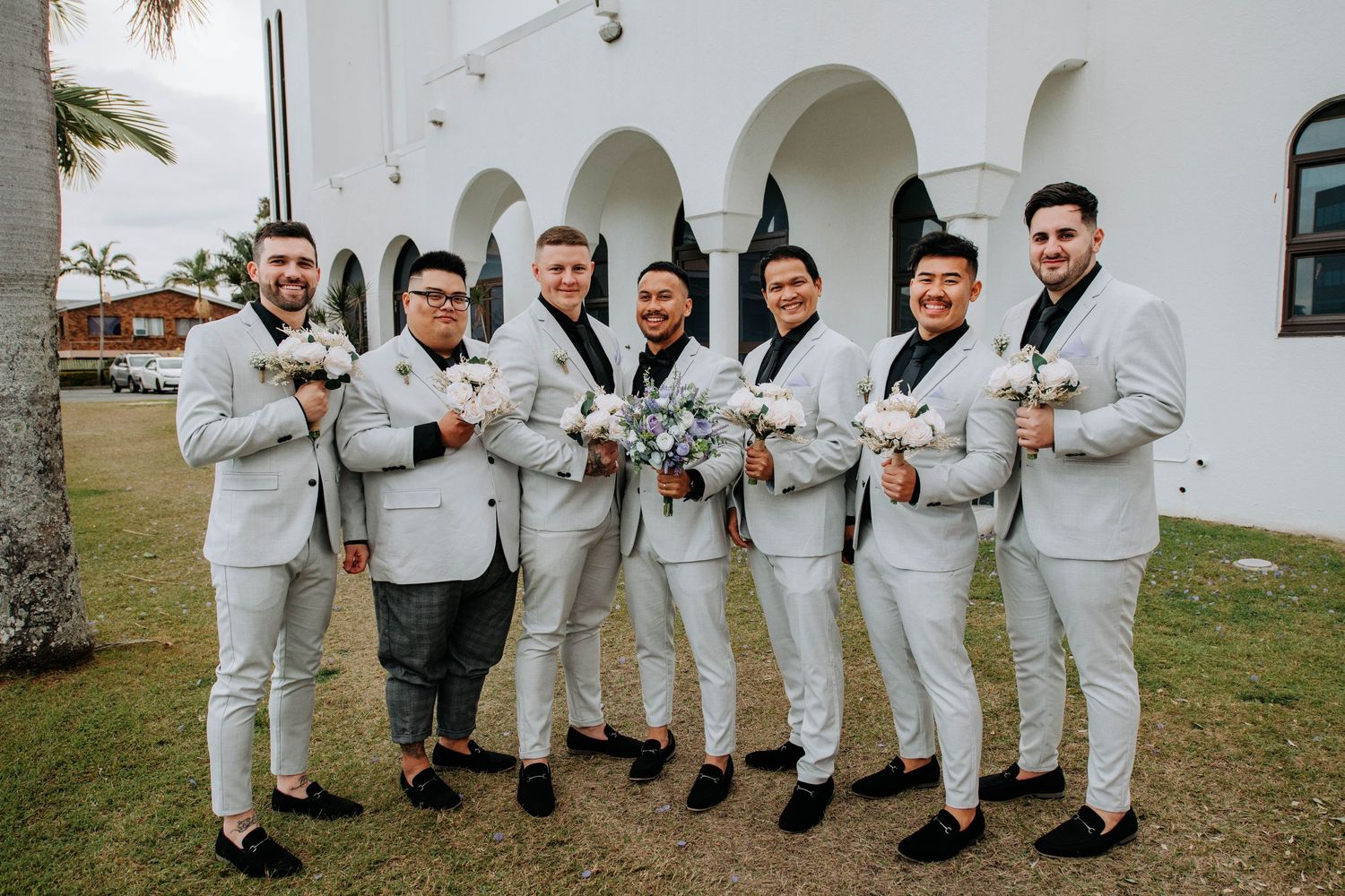 Groomsmen in light gray suits pose together near palm trees.
