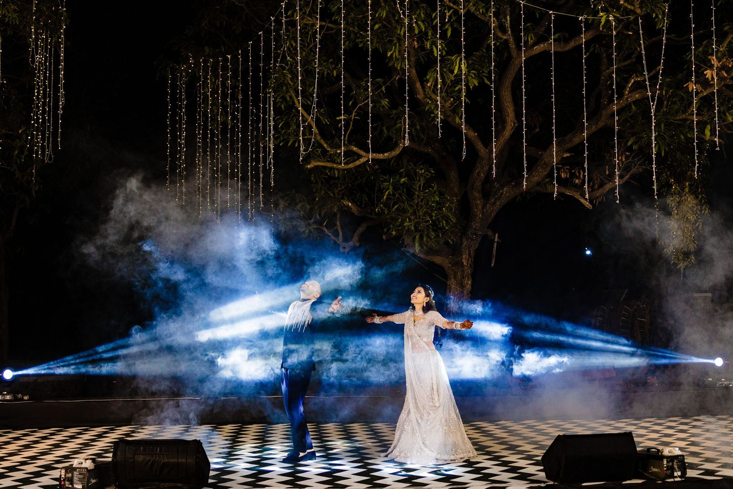 Dramatic nighttime wedding photoshoot with smoke effects and hanging string lights creating an ethereal atmosphere.
