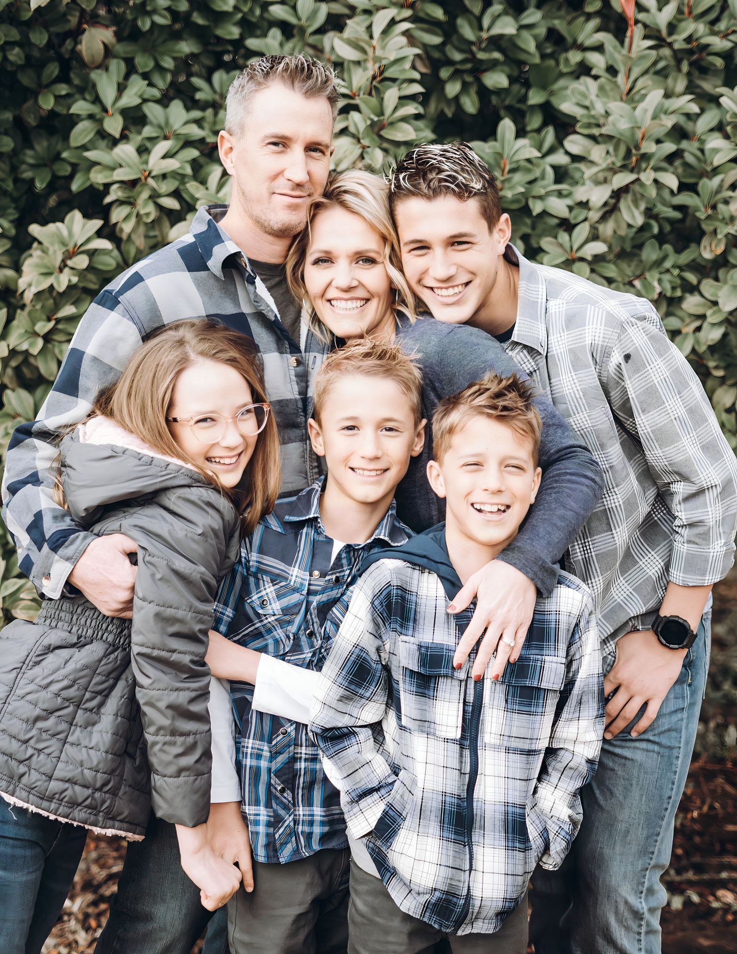 A happy family of six wearing plaid shirts poses together against a green leafy background.