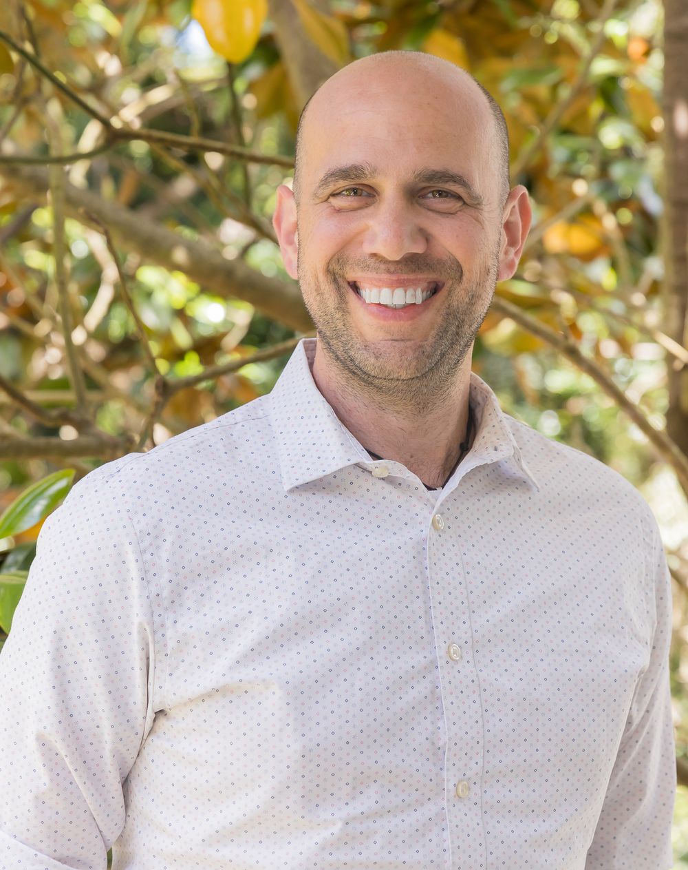 Professional headshot of person in white collared shirt smiling warmly against autumn foliage background.