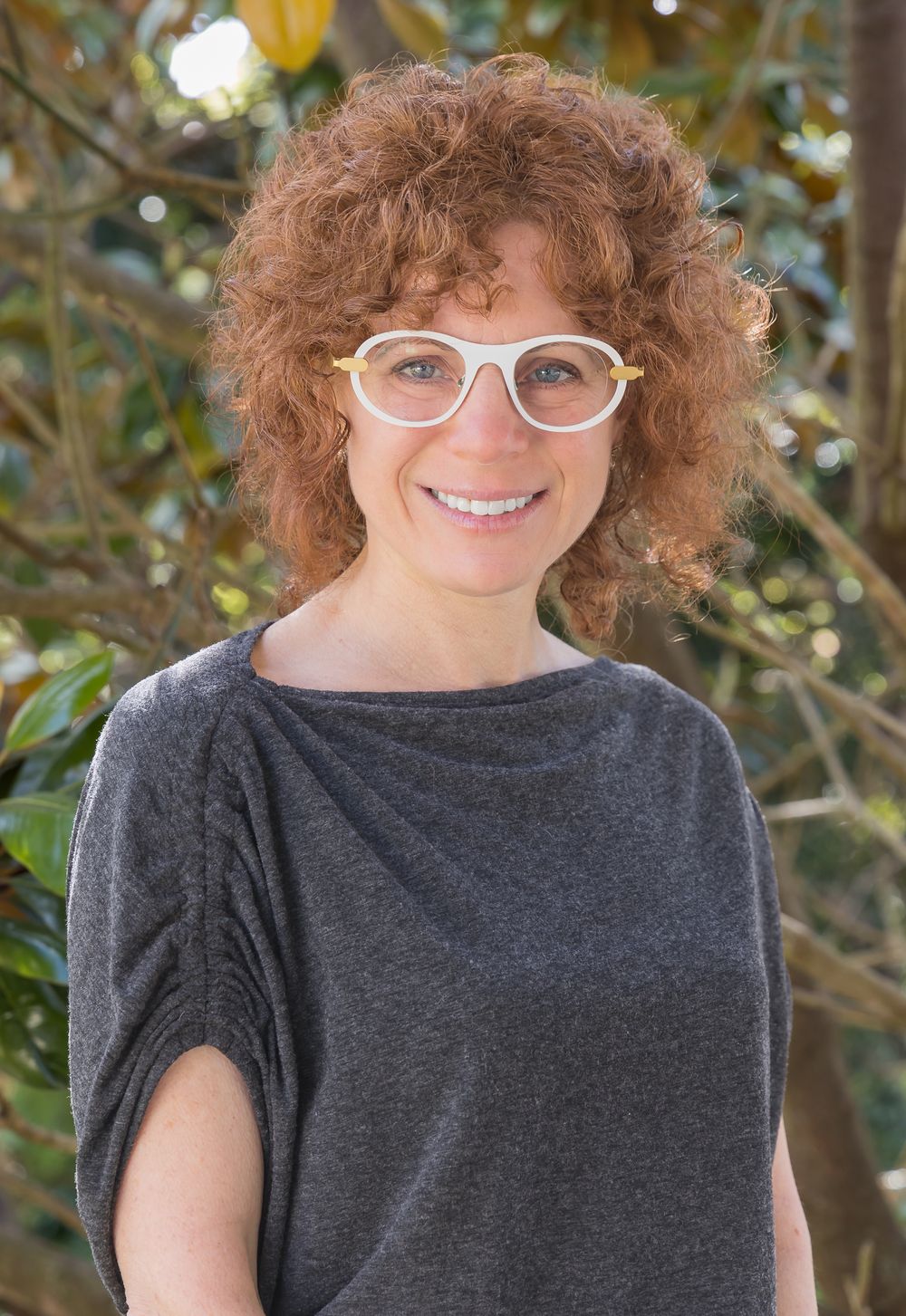 Portrait of person with curly hair wearing glasses and gray top against foliage.