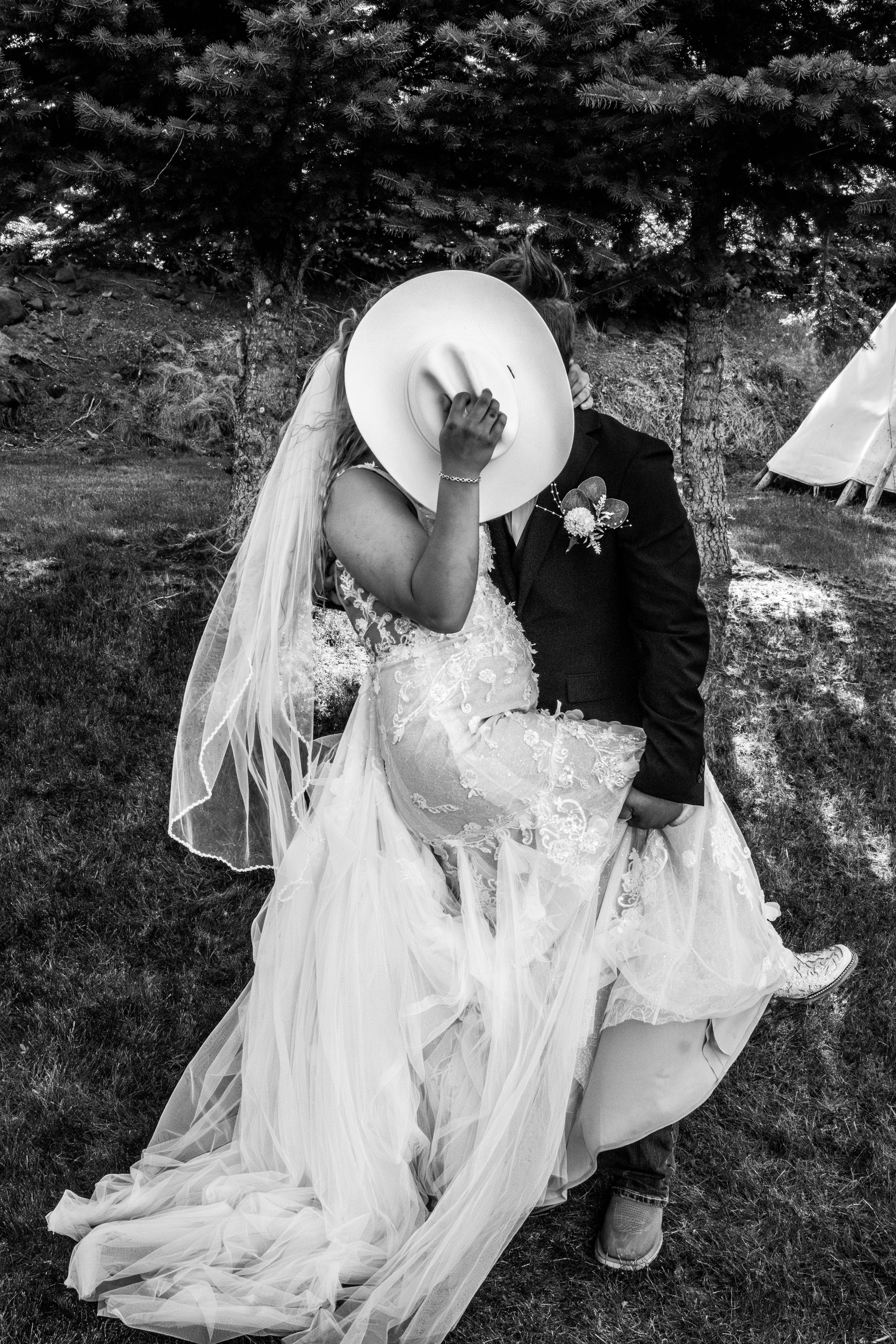A bride and groom share a kiss hidden behind a white cowboy hat during their outdoor wedding ceremony.
