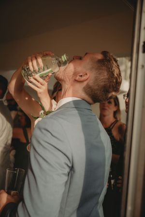 A wedding guest in a gray suit takes a celebratory drink from a bottle.