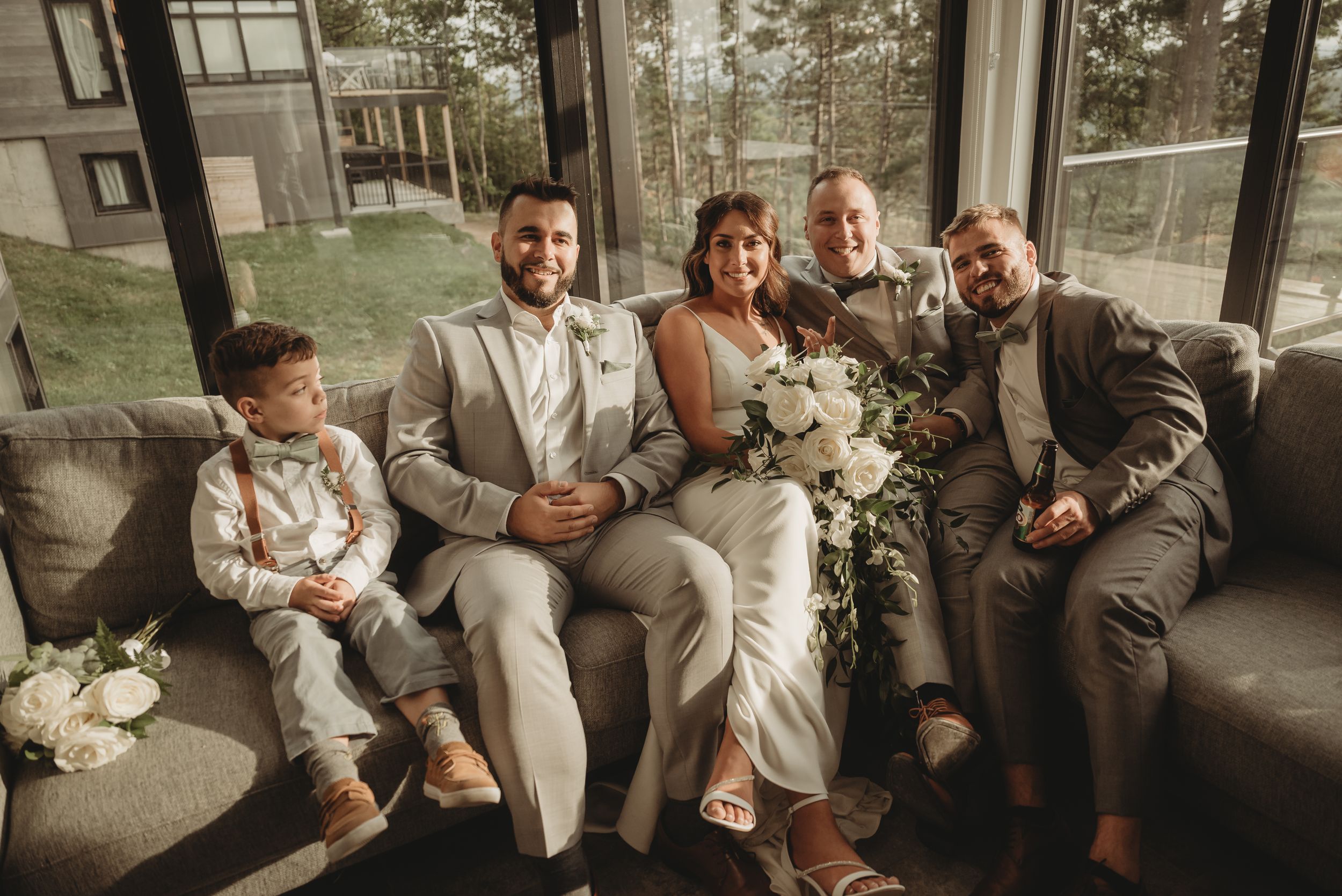A wedding party lounges on a gray couch near large windows with views of pine trees in the background.