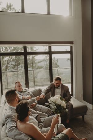 Wedding party relaxes on comfortable gray couches in a bright room with floor-to-ceiling windows.