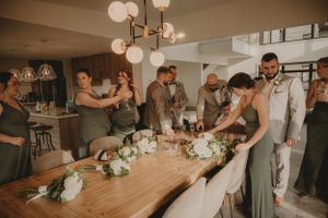 Wedding party gathers around a wooden table decorated with floral centerpieces in a modern venue.