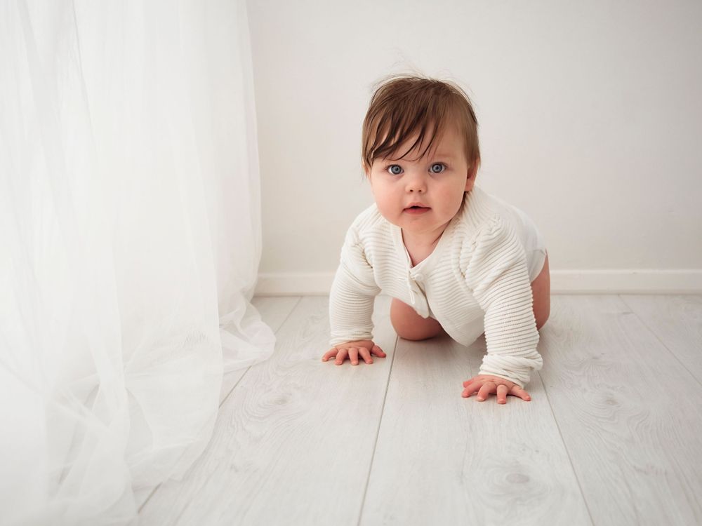 A 7 month baby in white jumper crawling beside a white curtain, Verity Photography