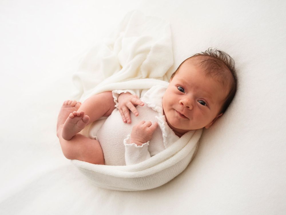 A newborn baby wrapped in a white blanket smiling during a photoshoot in Didcot, Oxfordshire, by Verity Photography