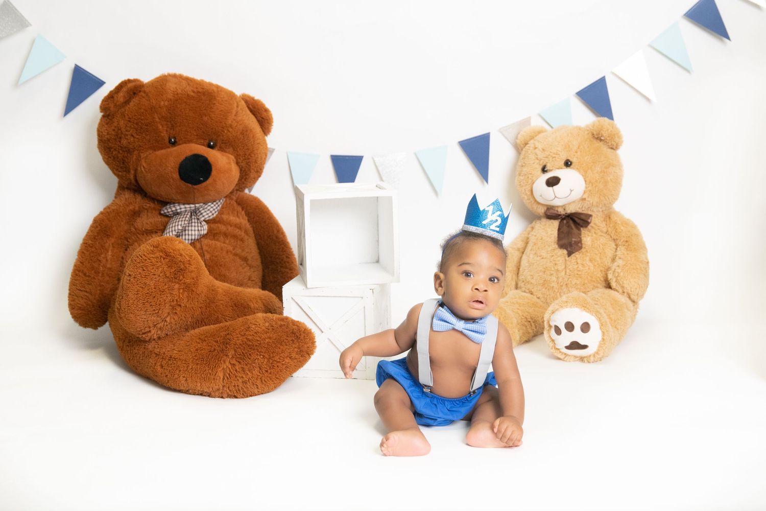 Baby photoshoot with teddy bears and blue banner decorations against white backdrop.