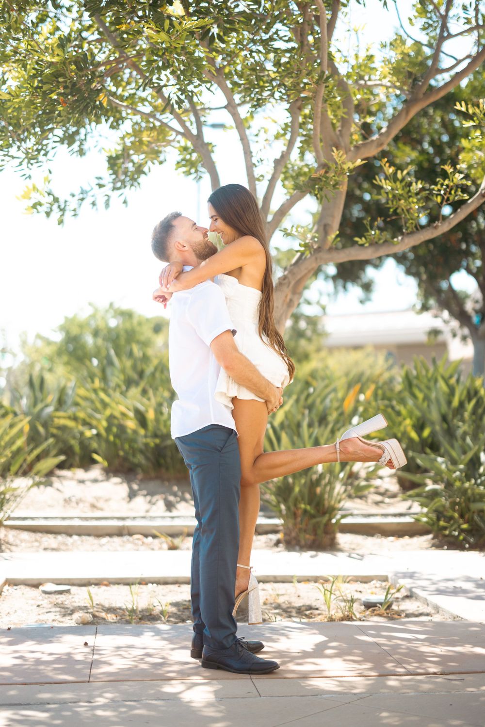 A romantic couple shares a playful lift moment in a garden setting with sunlit trees in the background.
