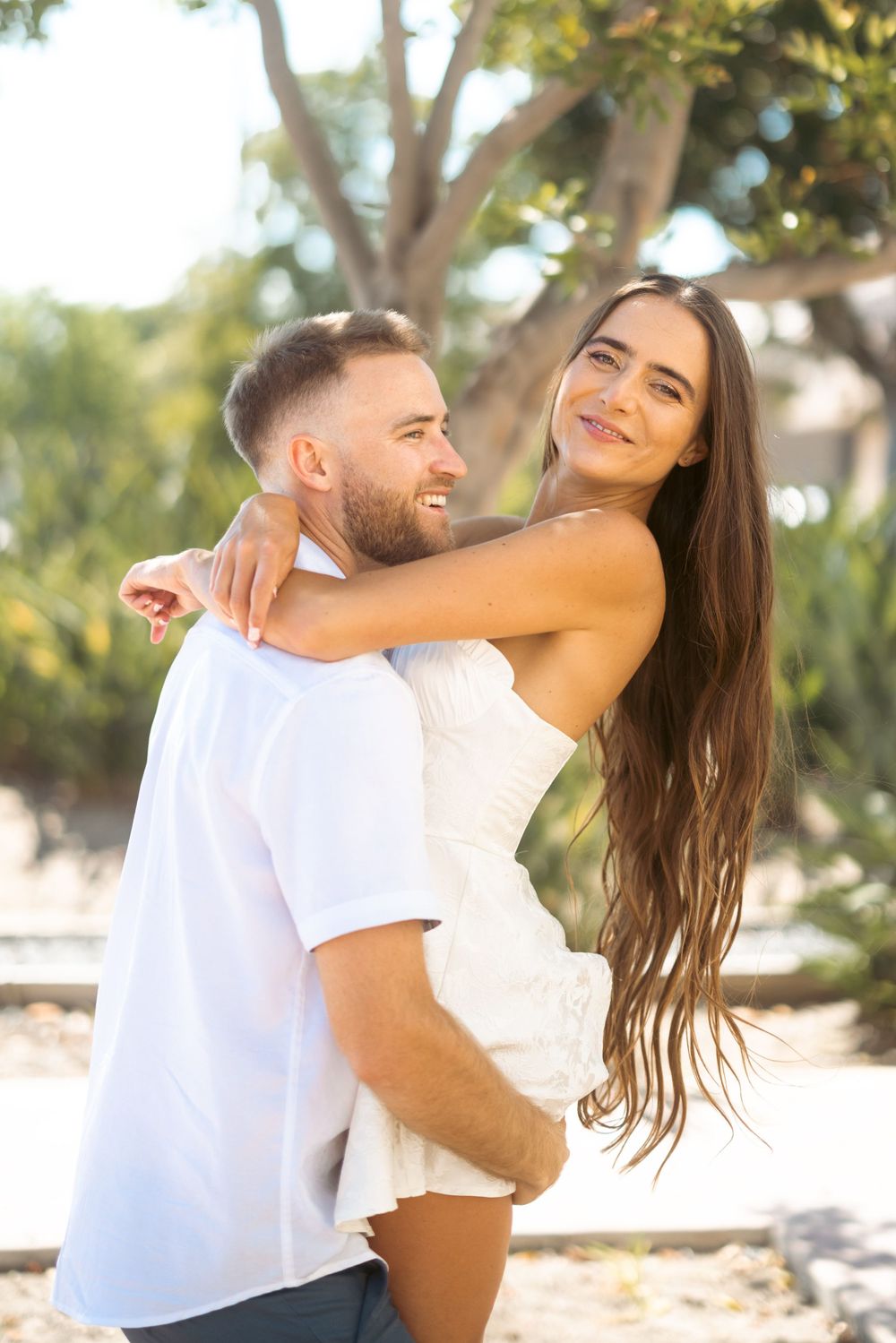 A happy couple in white outfits share a playful embrace in a sun-dappled garden.